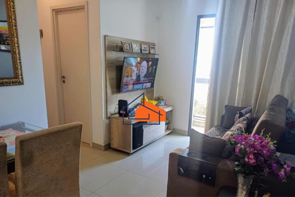 Sala de estar com sofá em L, TV de tela plana na parede, mesa com vaso de flores e cortinas brancas.
