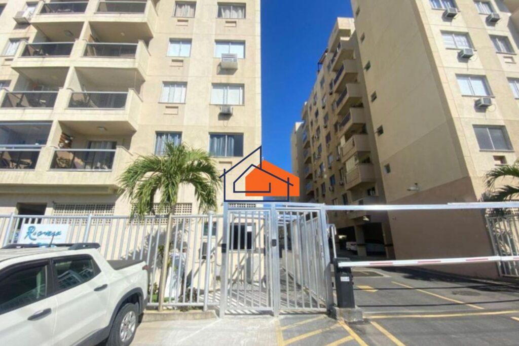 Entrada de condomínio residencial com portão metálico, guarita e prédios ao fundo.
