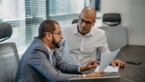 Dois homens em escritório analisando documento impresso sentados à mesa com persianas ao fundo.