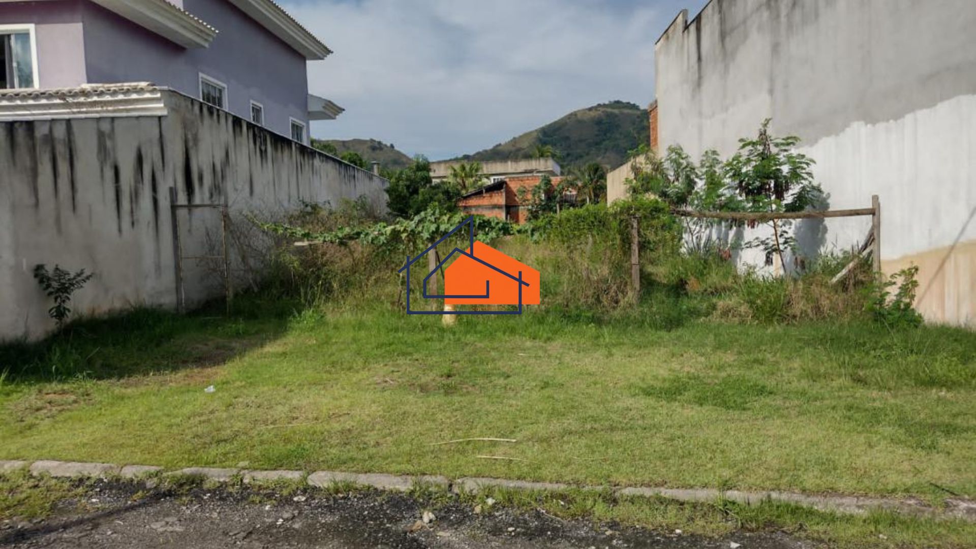 Terreno gramado vazio entre muros e casas, com vegetação ao fundo e montanha ao longe.