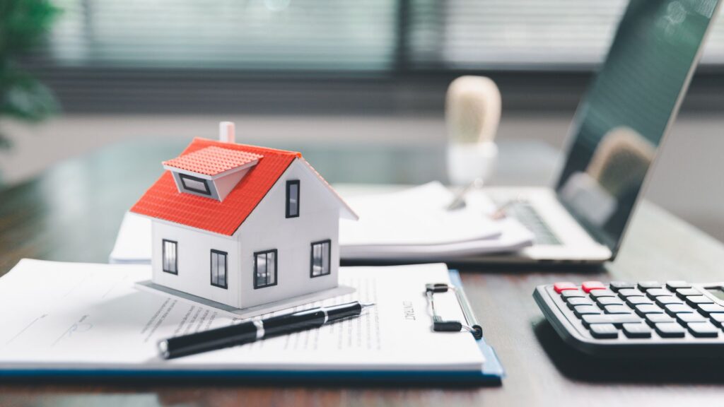 Modelo de casa sobre documentos com caneta, calculadora e laptop em mesa de escritório.