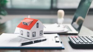 Modelo de casa sobre documentos com caneta, calculadora e laptop em mesa de escritório.